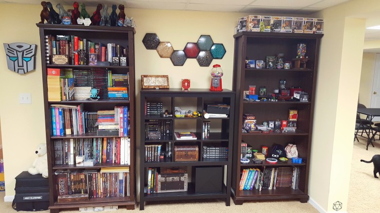 2 tall, wide bookcases flank a shorter shelf that has cube shelving. The right bookcase houses gamebooks and RPG books. The left bookcase houses dice, games with tin boxes, and other small gaming related trinkets. The central shelf houses gaming books, small chests with games inside, and the Invisible Sun Black Cube. Above the central shelf is a metal art piece (a series of hexagons in dark colors). 